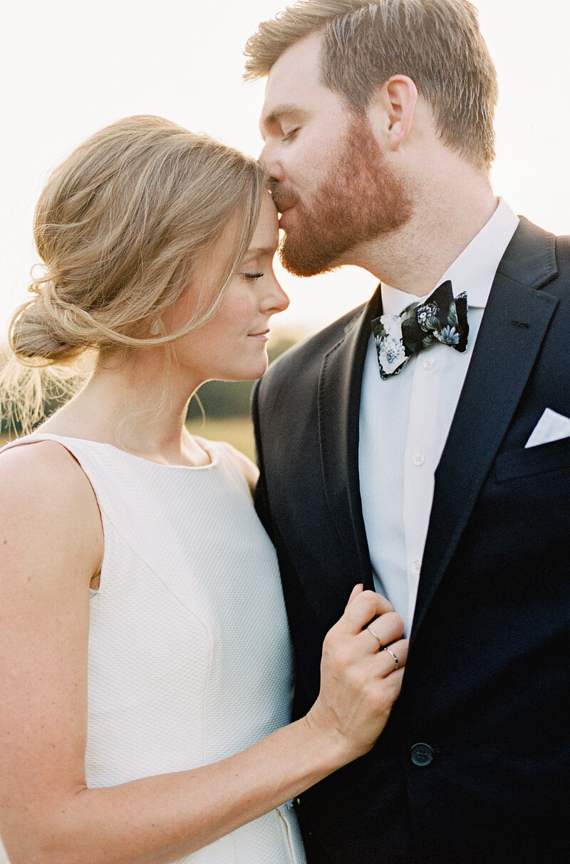 A groom with a subtle floral bow tie tenderly kiss the forehead of a bride in an elegant dress as he passionately grips his lapel. | Wedding Photographer in Pittsburgh PA | Anna Laero Photography