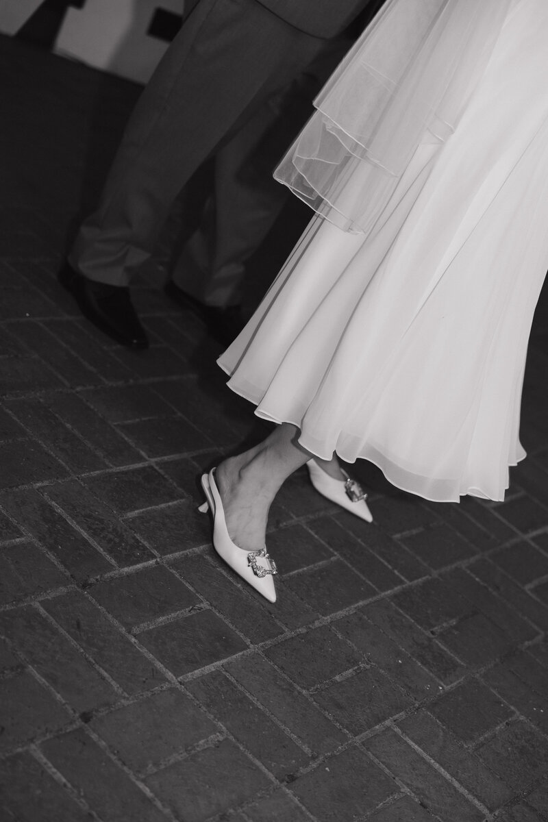 Black and white photo of bride’s pointed toe heels and the hem of her wedding dress on a brick walkway.
