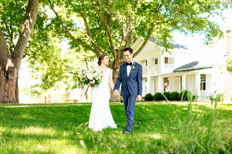 A wedding couple walking and holding hands in front of the white farm house at Zingermans Cornman Farms in Dexter Michigan