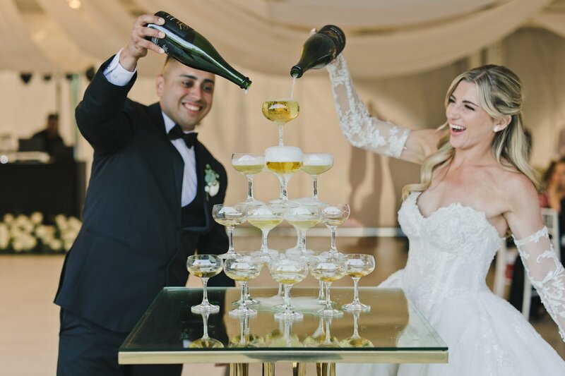 A bridge and groom smiling as they pour champagne over a tower of glasses in a documentary style wedding photo taken by One Love Photography.
