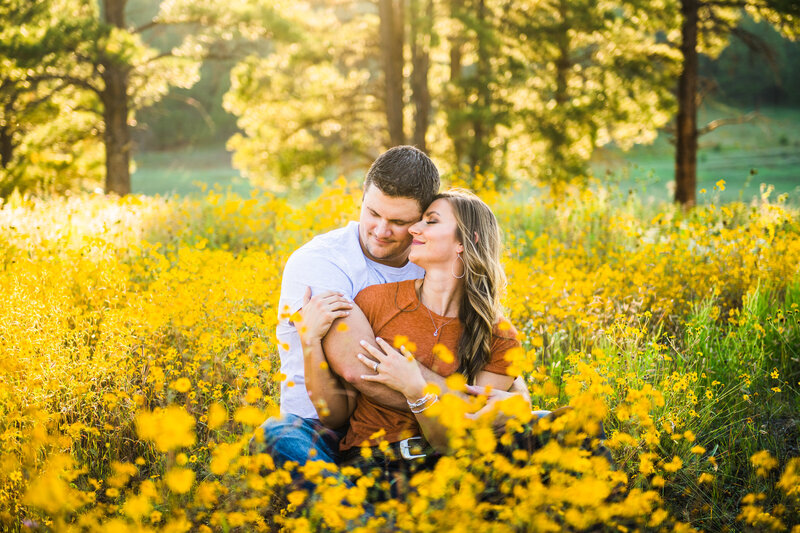 Engaged couple holding each other sitting down with eyes closed in yellow wild flower field  and pine trees at golden hour in Flagstaff Arizona 