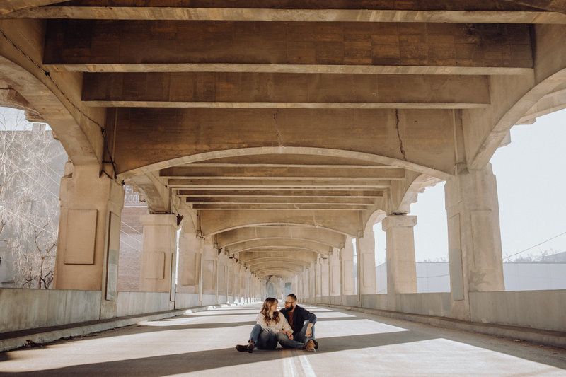 Couple sitting side by side in the middle of the road on a bridge, looking at each other, smiling. Location is in the West Bottoms in Kansas City, MO.