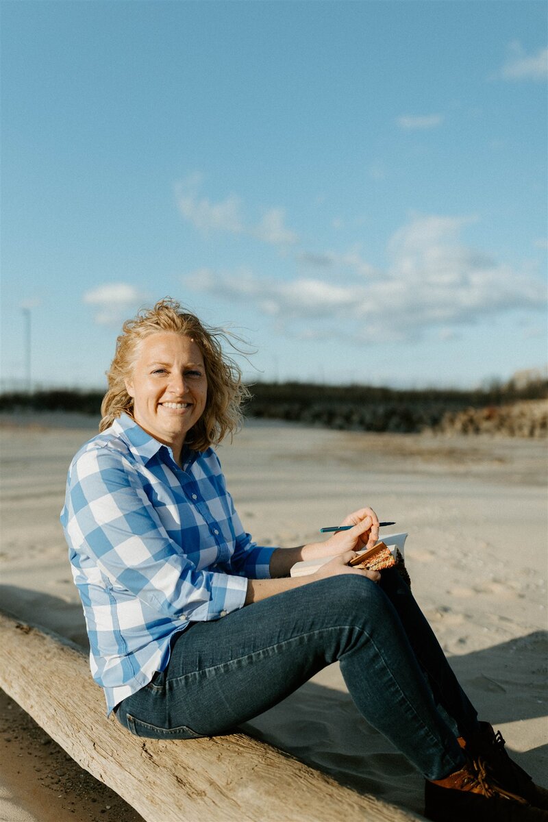 Writer, Sarah Freymuth, smiles for photo in black v-neck t-shirt while sitting on tree roots outdoors