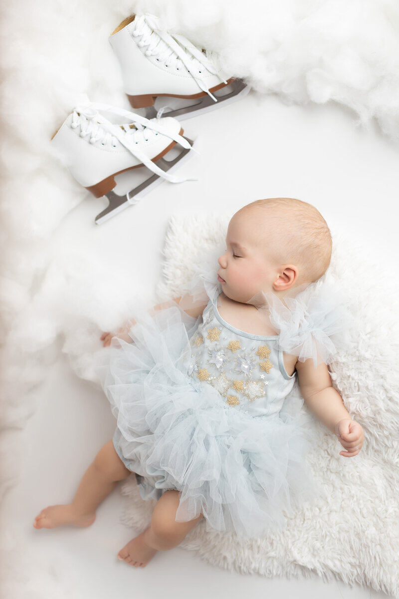 Baby girl sleeps peacefully in a soft blue tulle dress with ice skates nearby, surrounded by fluffy clouds and white textures. This winter-themed milestone portrait was captured by Dallas Baby Milestone Photographer Lindsay Walden.