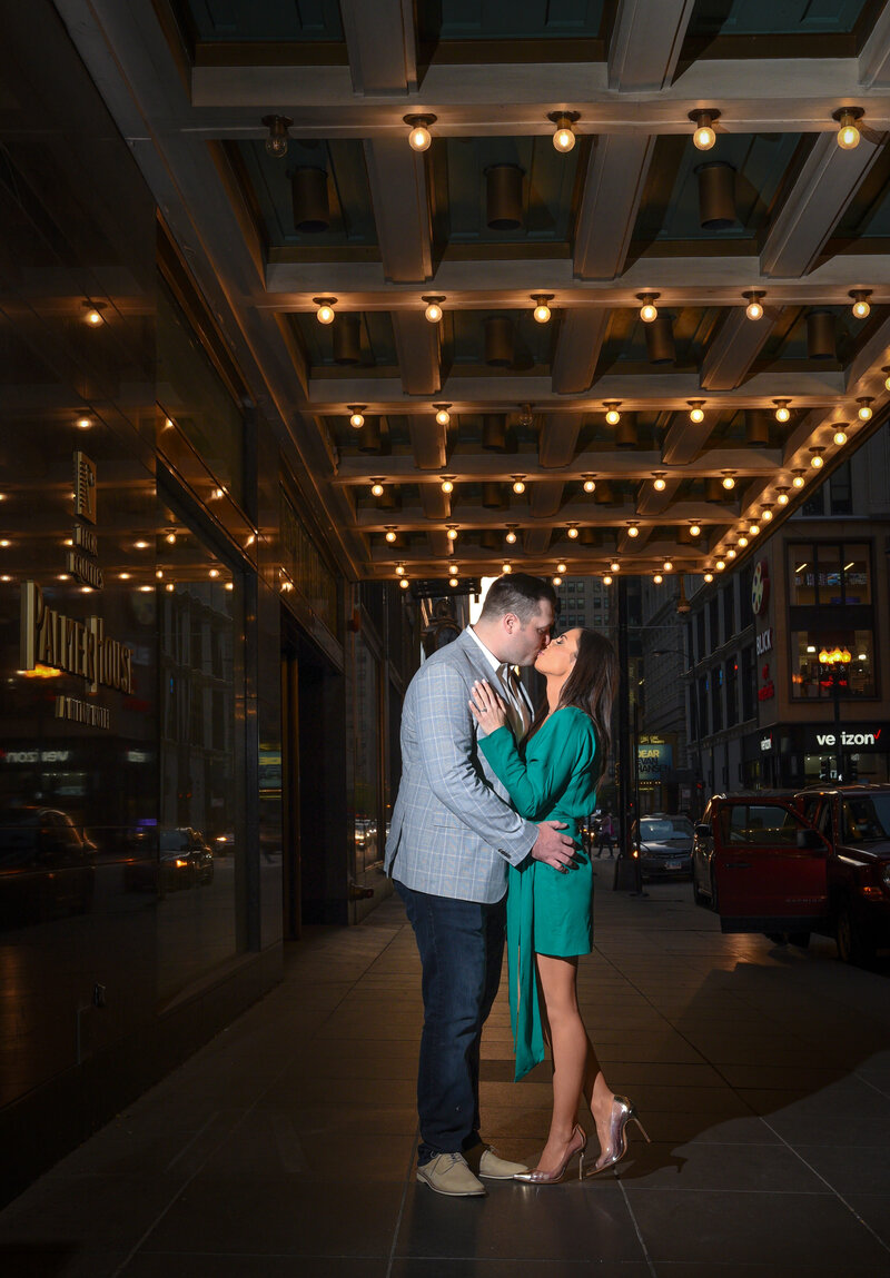 Woman in green dress kisses her fiance underneath a lit marquee