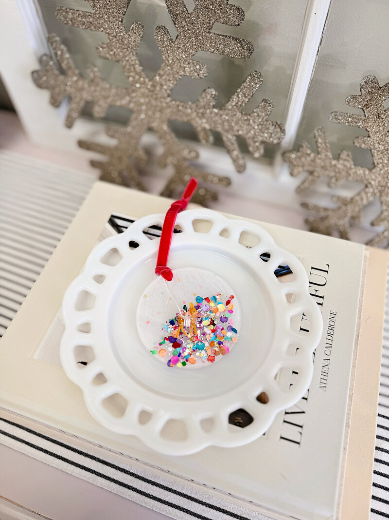 A round white holiday ornament from Sweet Pea Ink Creative with colorful confetti, a tiny reindeer, and a red ribbon rests on a decorative white plate atop a book, with glittery snowflake decor in the background.
