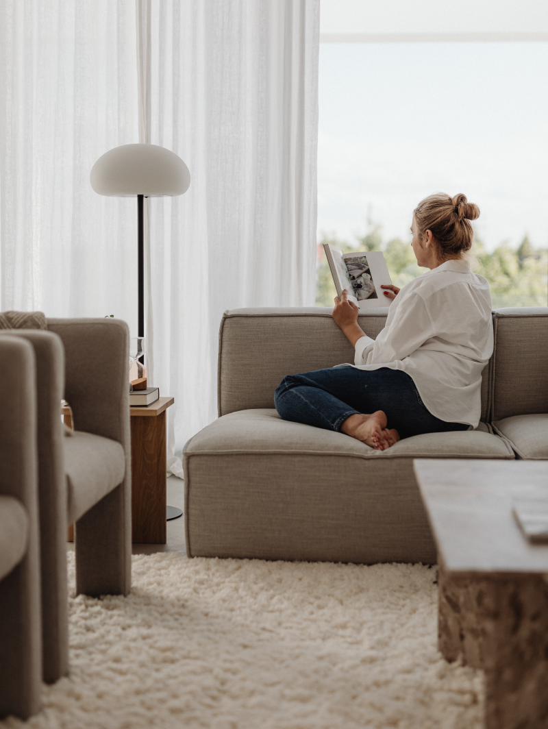 Young woman sitting on a couch reading a book in a comfortable apartment |Brand By Design