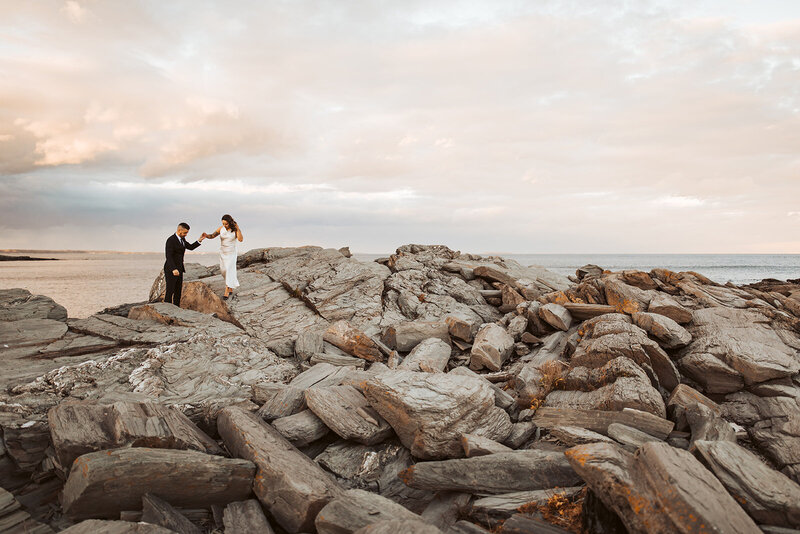 Bride and groom holding hands and walking on the rocks on the coast of maine at sunset.