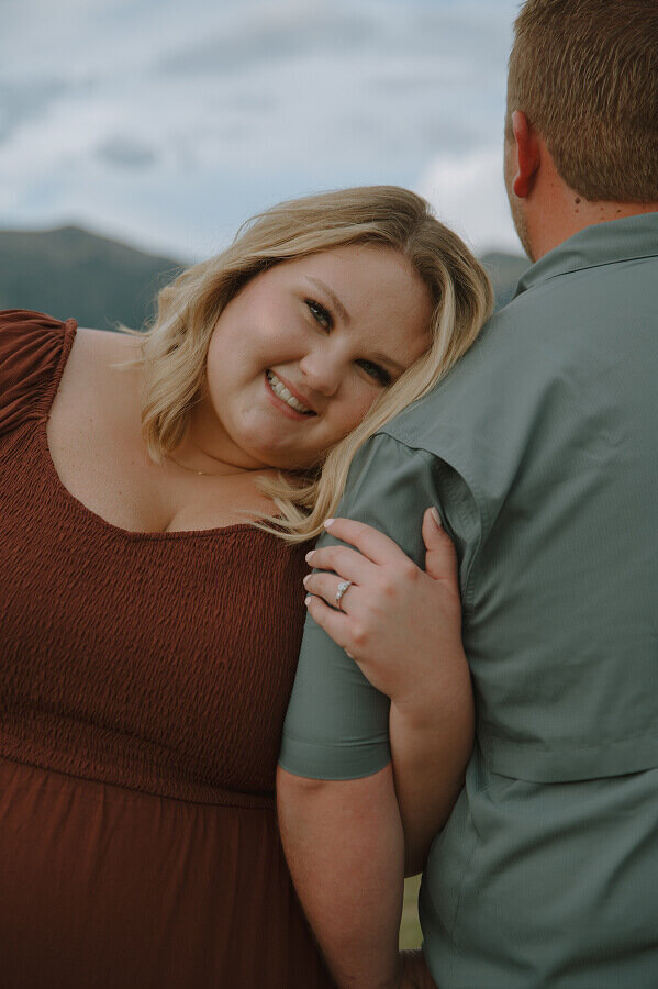 Close up of a woman smiling at the camera. She is resting her head on her fiancé's shoulder and holding his arm. 