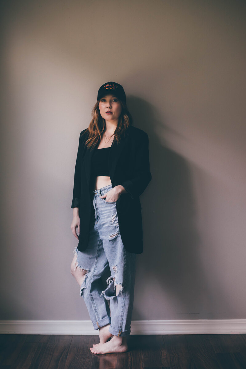 A person stands against a wall, wearing a black blazer over a black top, distressed jeans, and a baseball cap. Their arms are relaxed by their sides, and they are barefoot—embodying the casual yet confident look of a web designer for small business projects.