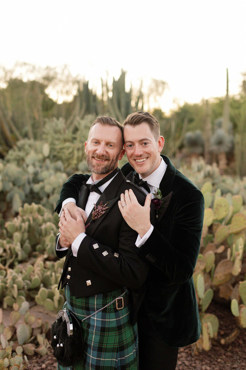 Two grooms cuddle and smile in a cactus garden. 