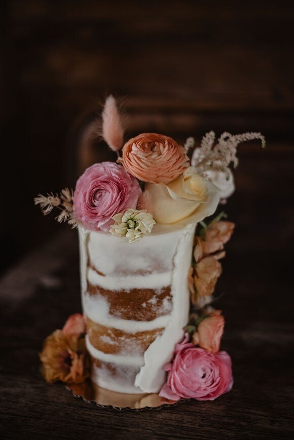Photo shows a small cake, with naked style frosting (some cake showing through the frosting), and then a partial wrap of white buttercream with a rough edge around the side. Cake is decorated with a large assortment of flowers in pinks, oranges, and yellows with some on top, some going down the side, and a couple at the bottom. Flowers are mainly ranunculus and roses.