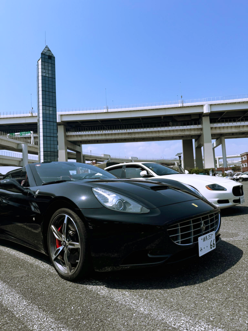 A black Ferrari California and a white Maserati Quattroporte parked side by side under the Tokyo expressway at Daikoku Parking Area, ready for a luxury drive experience.