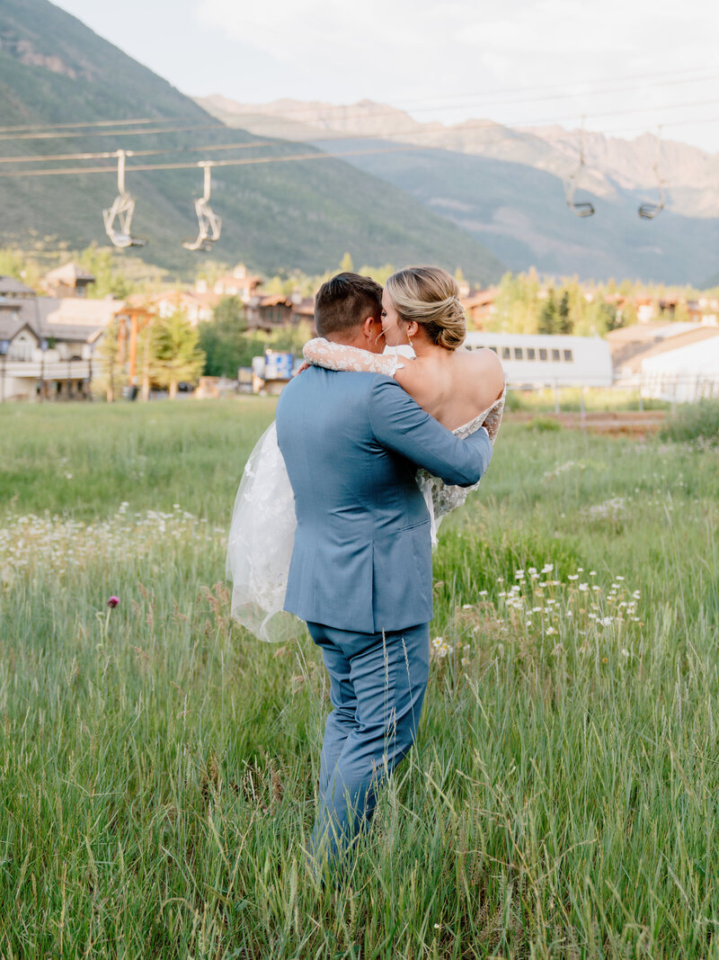 A groom carrying his bride in a field of tall grass, in the background there are mountains and ski lifts