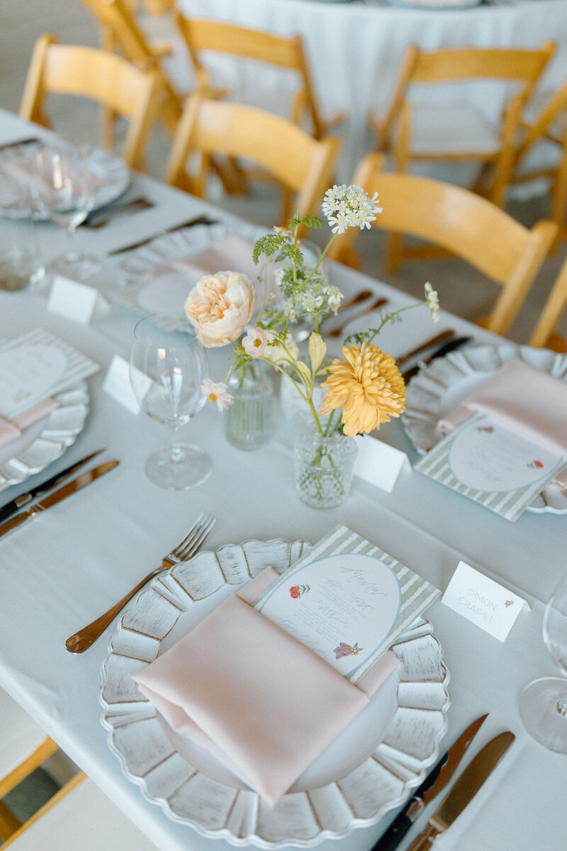 A wedding reception table is set for guests with flowers and place settings featuring name cards and menus with customized calligraphy.