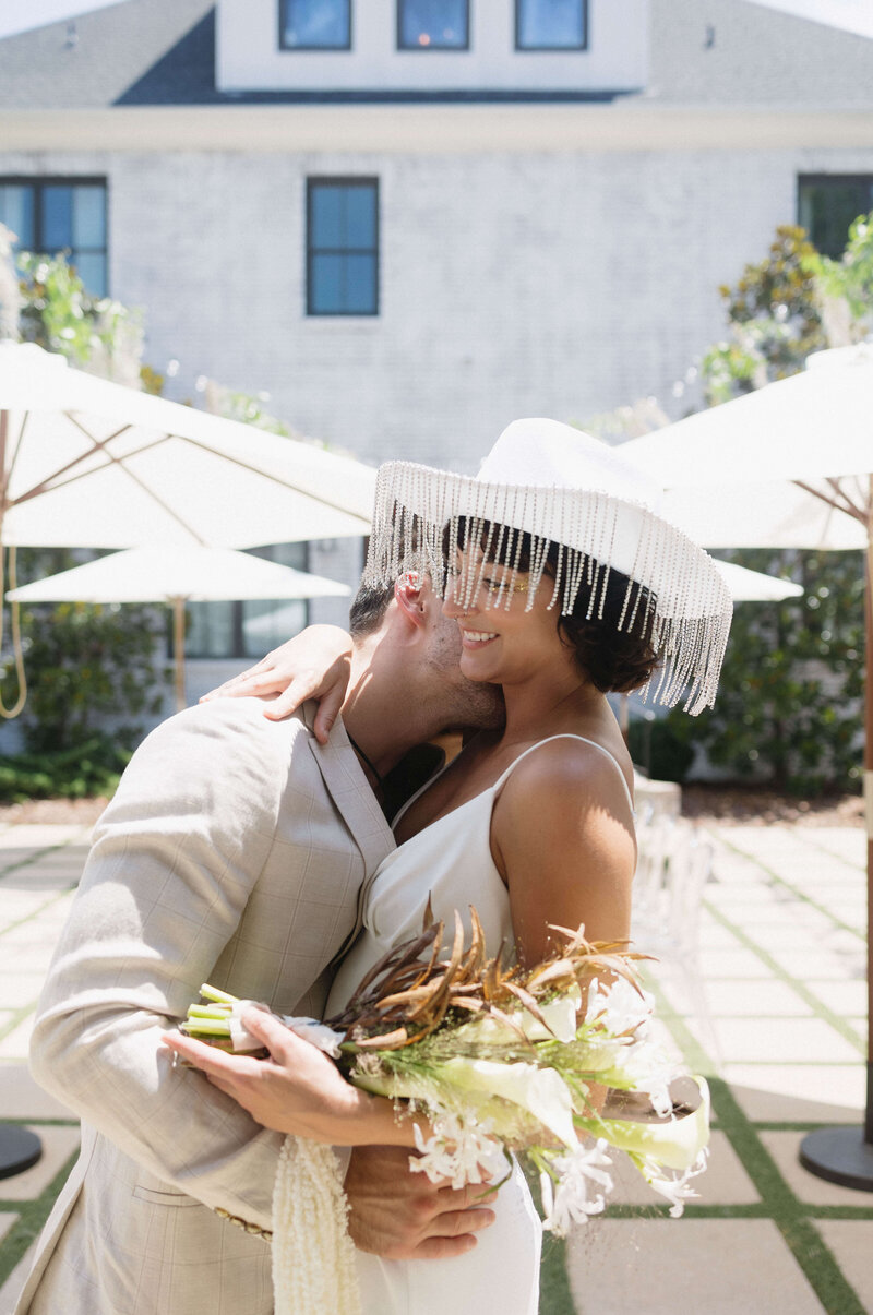 The groom sweetly kisses the bride's neck as she holds a beautiful wedding bouquet and wears a charming hat
