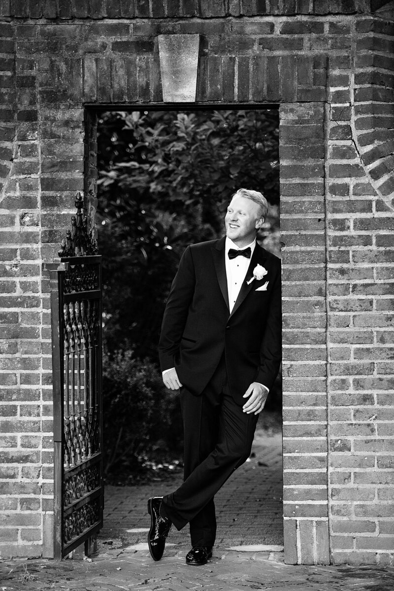 A black and white image of the groom leans in the door way of a stone entrance with a decorative iron gate. there are magnolia trees behind him. He is wearing a black tuxedo with a white shirt and a black bowtie. He has a white boutonniere pinned to his lapel.