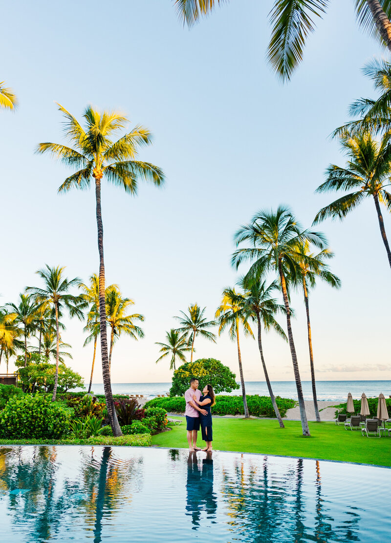 couple stands at the edge of the  resort pool  with blue water and palm trees