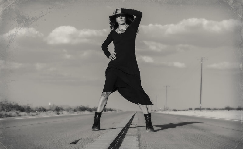 Musician portrait Johnette Napolitano standing in middle of road arm up covering hat brim black and white