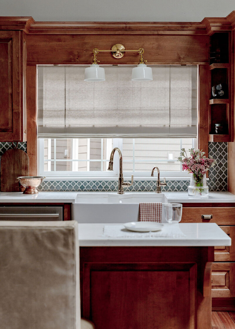 A kitchen farmhouse sink under a window, with a backsplash and wooden cabinets.