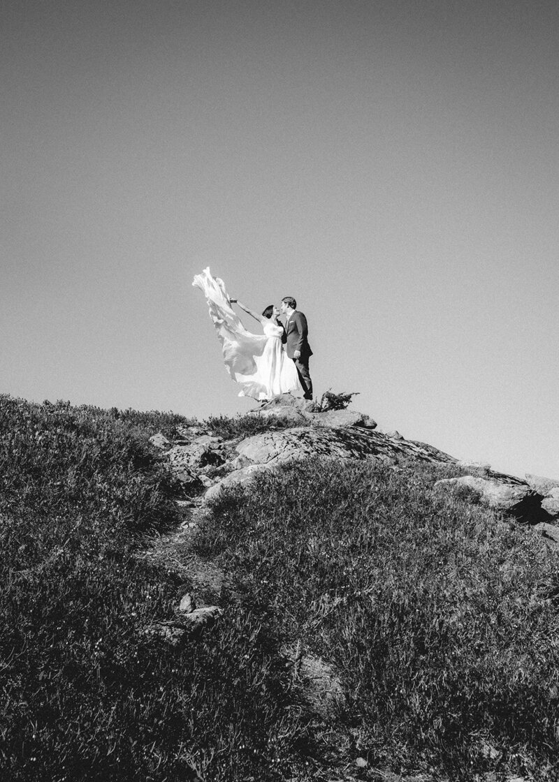 from a full day elopement package, a bride and groom in their wedding attire share a kiss at the top of a hill. A dirt path creates a leading line as the bride flings her dress to the wind. The lighting is harsh and overhead having passed the morning golden hour, and creates a dramatic effect. 