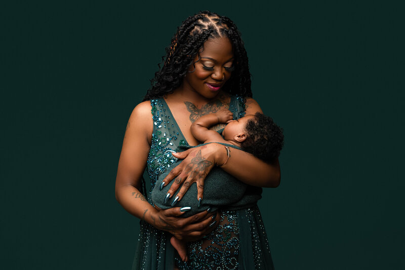 Mother lovingly breastfeeding her baby while wearing a green sequined gown against a deep green backdrop.