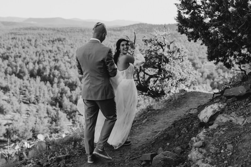 bride looking back at groom helping her with her dress on their hike to take elopement wedding portraits