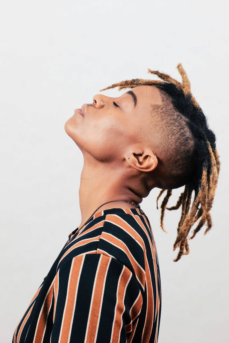This image shows a young person of color with dreadlocks in profile, lifting their head back, face toward the ceiling with closed eyes.