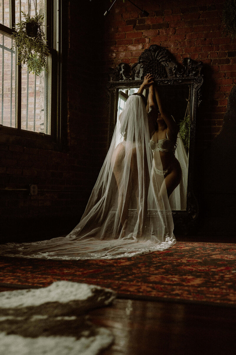 Bridal boudoir photo of a woman in sheer white lingerie and a cathedral veil, standing in front of an ornate mirror in a moody loft studio with brick walls and vintage decor