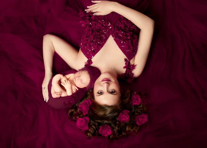 Newborn photography session — mother in burgundy gown cradling baby wrapped in matching swaddle, styled with rose accents for studio portrait