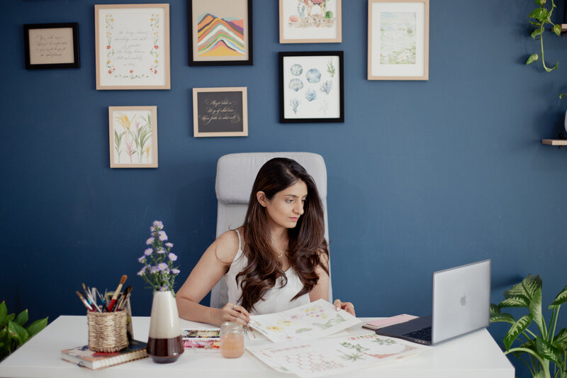 Komal, an Oman-based artist, works from her home studio, flipping through her designs while working on her laptop. She specializes in creating bespoke wedding stationery and watercolor illustrations for clients worldwide.