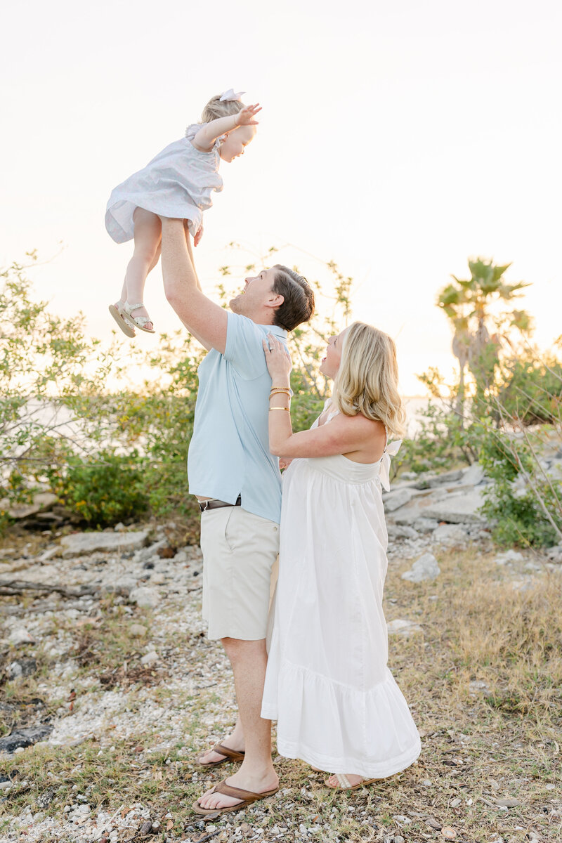 Dad throwing toddler daughter up in the air while pregnant mother stands behind and smiles in white maternity dress outside in Tampa, Florida