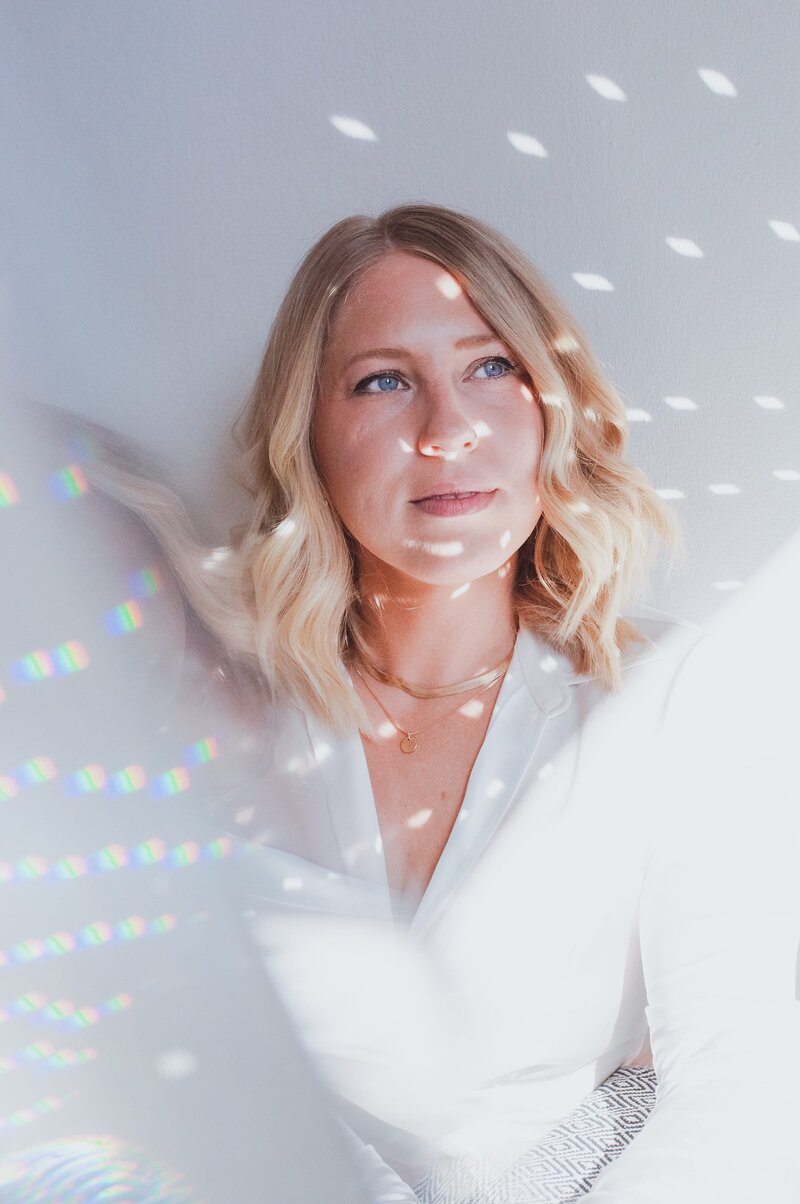 Woman with blonde, wavy hair and silk, white top with disco ball reflections on her face as she looks away.