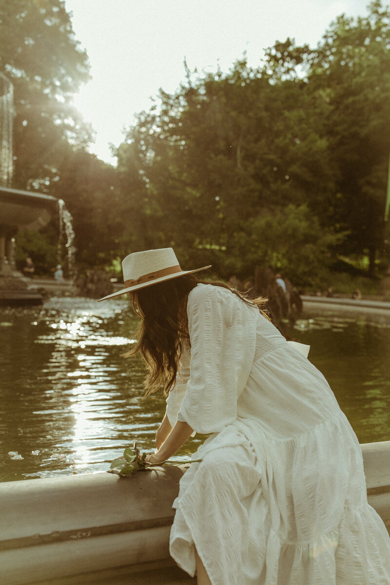 Woman in  a white dress and straw hat with flowers in her hand leaning over a fountain in the park at sunset