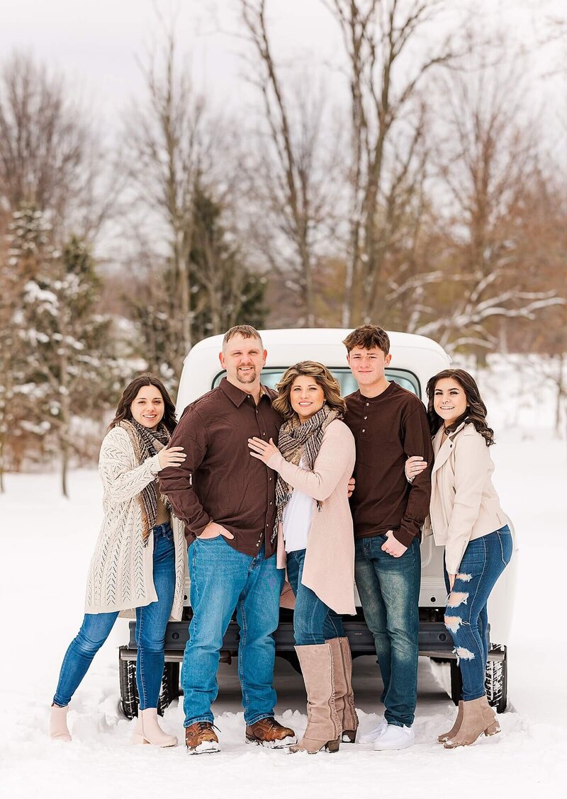 Family with beige, tan, and light pink outfits standing by a white truck in the snow for a family portrait session.
