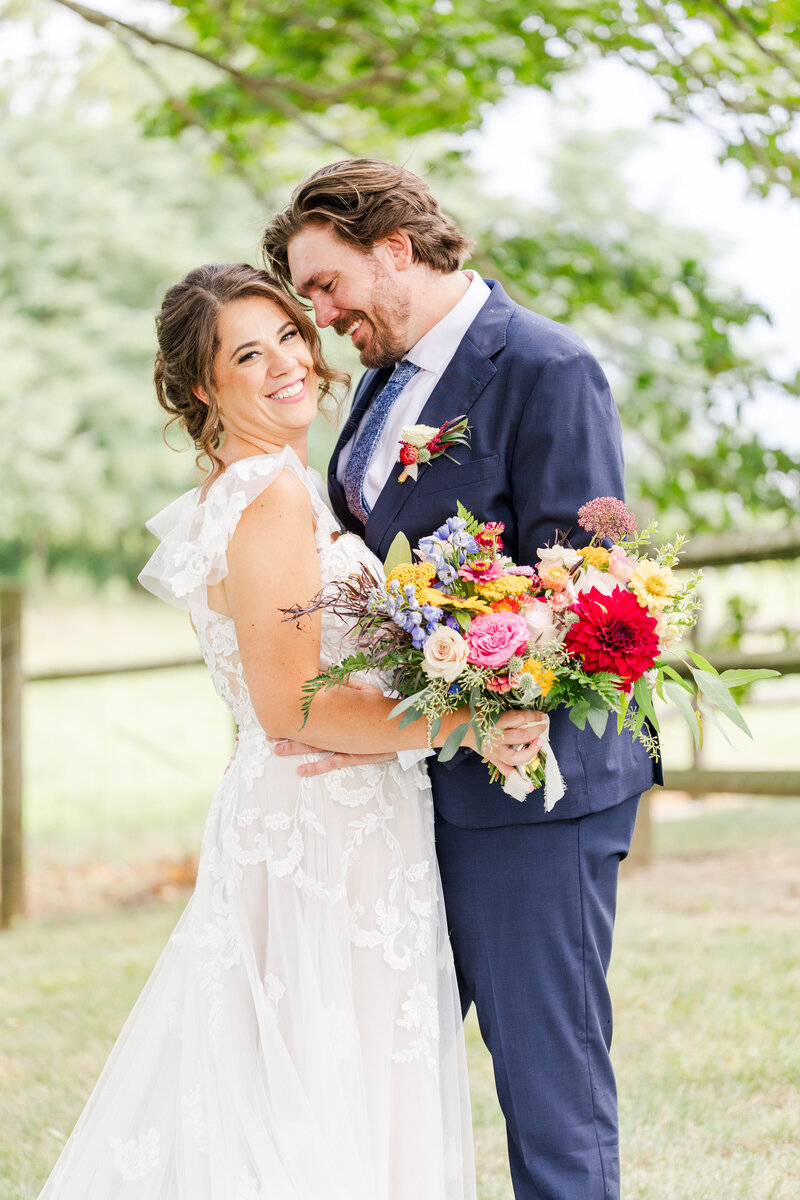 Newlyweds dance in a field in a blue suit and lace dress holding a colorful bouquet and laughing for a Detroit Wedding Photographer