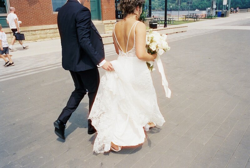 bride and groom walking together on wedding day while bride holds bouquet and groom holds back of dress by minnesota photographer tom thornton photography
