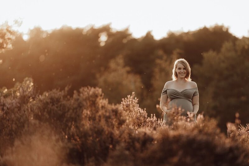 Zwangerschapsfoto van een zwangere vrouw tussen heidebloemen bij zonsondergang, warme natuuromgeving en dromerige sfeer.