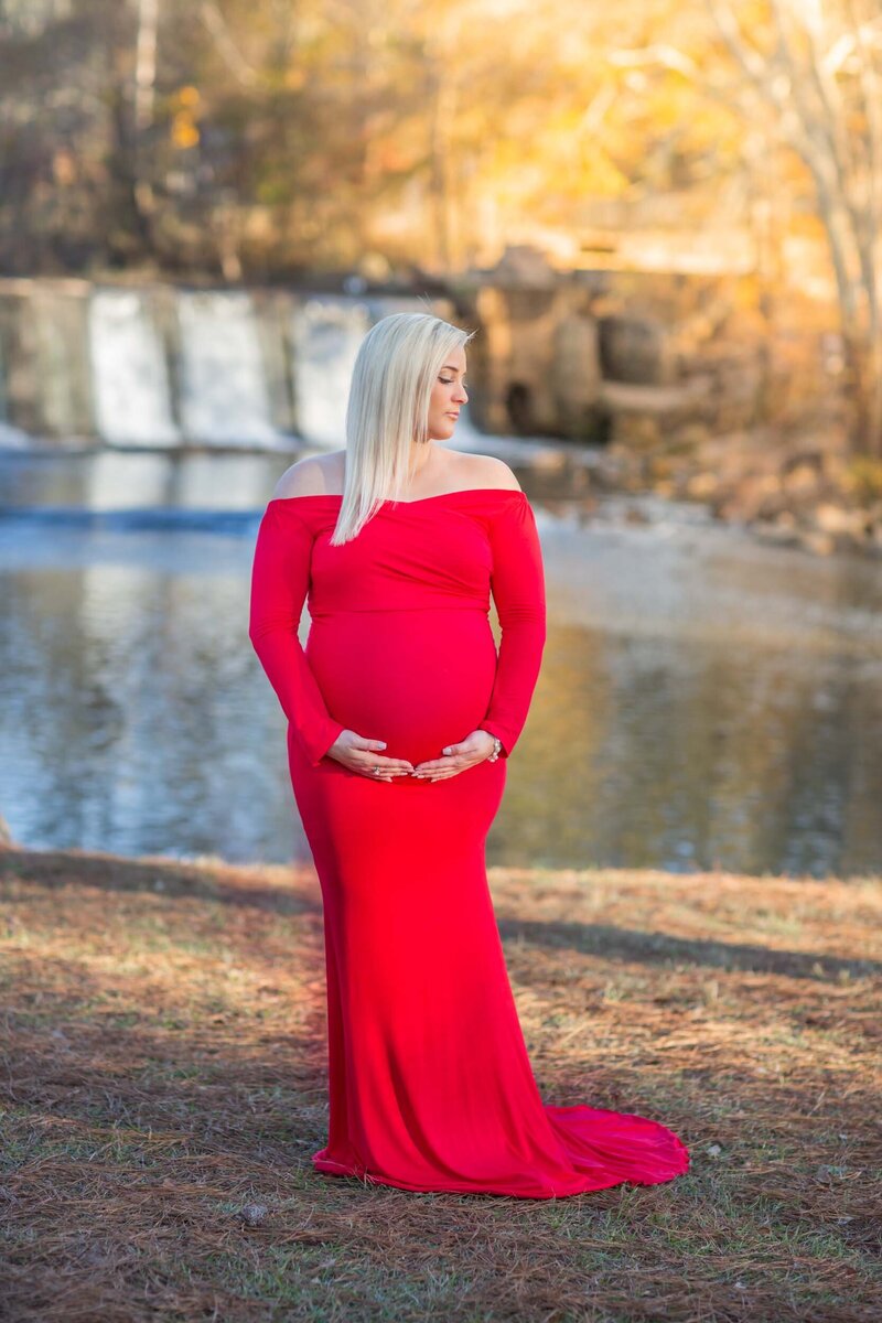 An expecting mother in a bright red, flowing dress holds her belly lovingly while looking into the distance at Starr's Mill Waterfall.