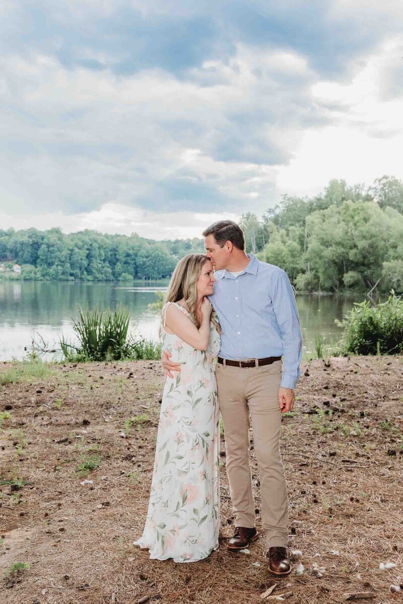 A man pulls his woman in tight for a kiss on the forehead in front of Lake Kedron in Peachtree City during their engagement portraits.