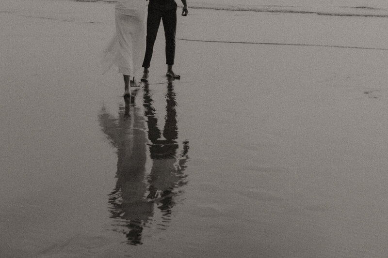 Back and white detail photo of a couple walking along the beach holding hands. Photo is of their waist down with their full bodies reflected in the wet sand.