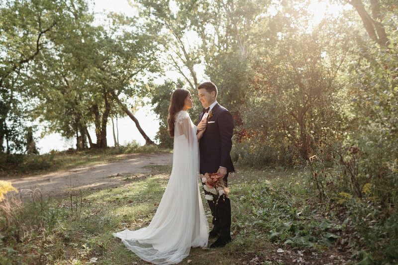 In a forested haven, with sunlight streaming through the trees, a bride and groom stand facing each other in formal elegance. Captured beautifully by a skilled Toronto wedding photographer, the moment is timeless as she holds her bouquet, surrounded by nature’s serene embrace.