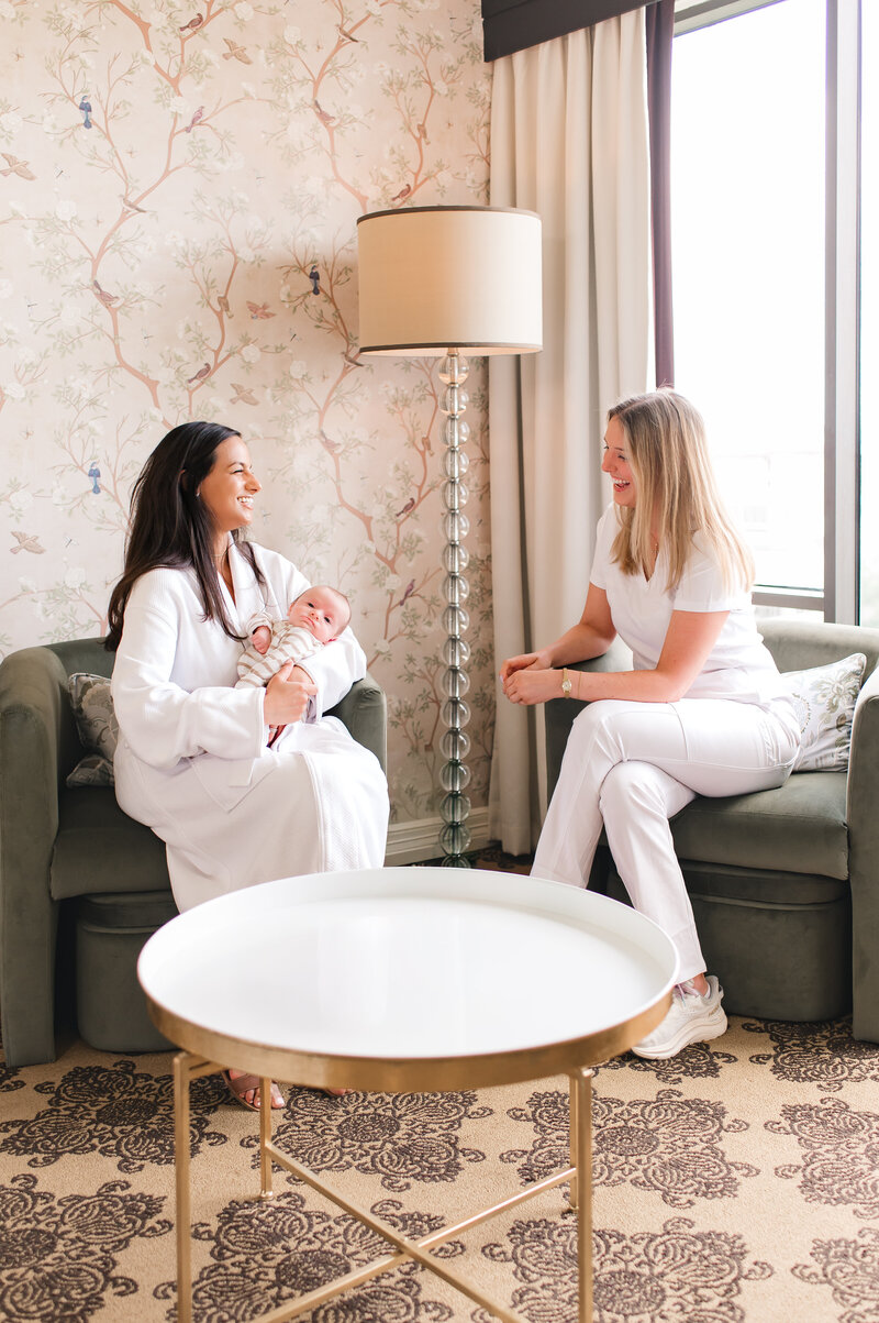 New mother holding her newborn while consulting with a postpartum nurse in a hotel suite at The Ruby Postnatal Retreat in Houston