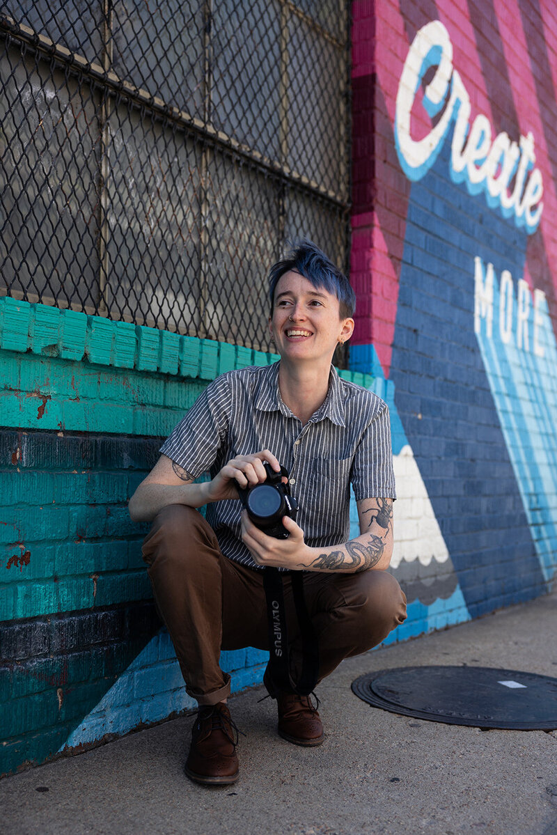 photographer River Dantzler smiles and holds up their camera. they have short blue hair and tattoos and wear a striped button down shirt.