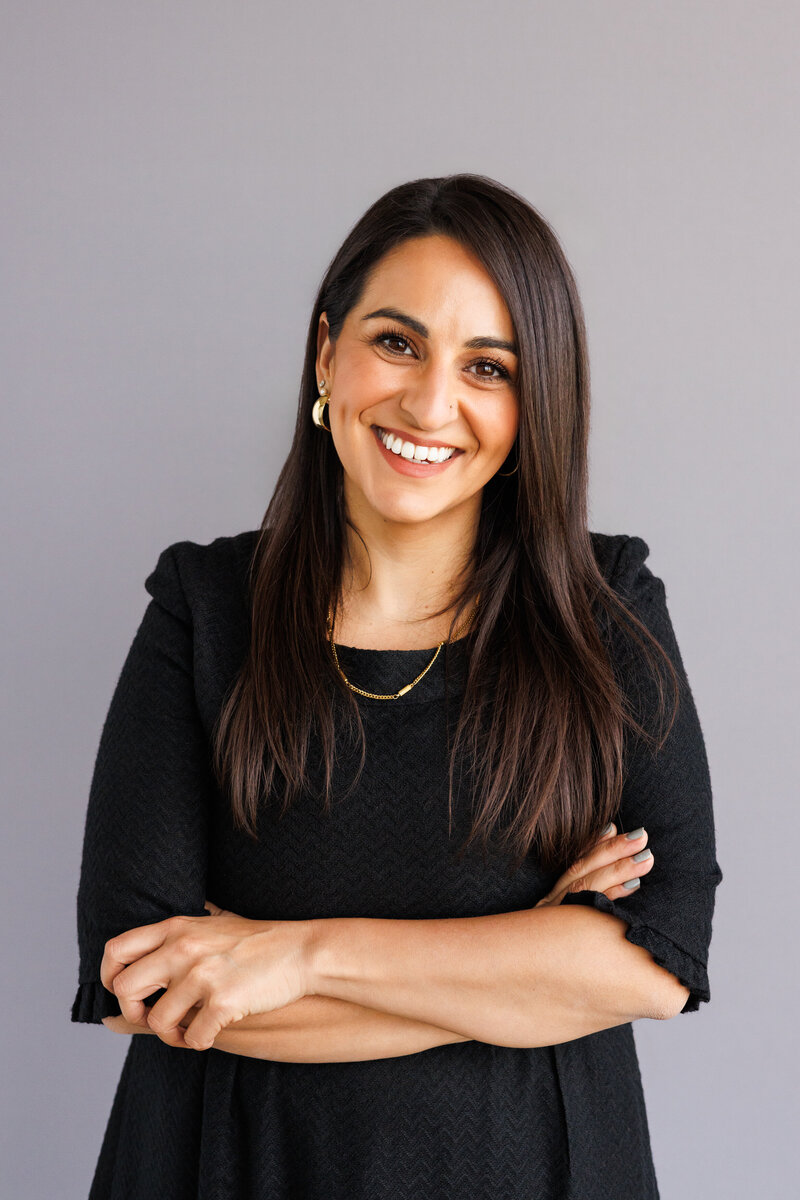 A woman with long, dark hair smiles warmly with her arms crossed, wearing a black textured dress and gold jewelry against a neutral grey background. A polished and approachable professional headshot.