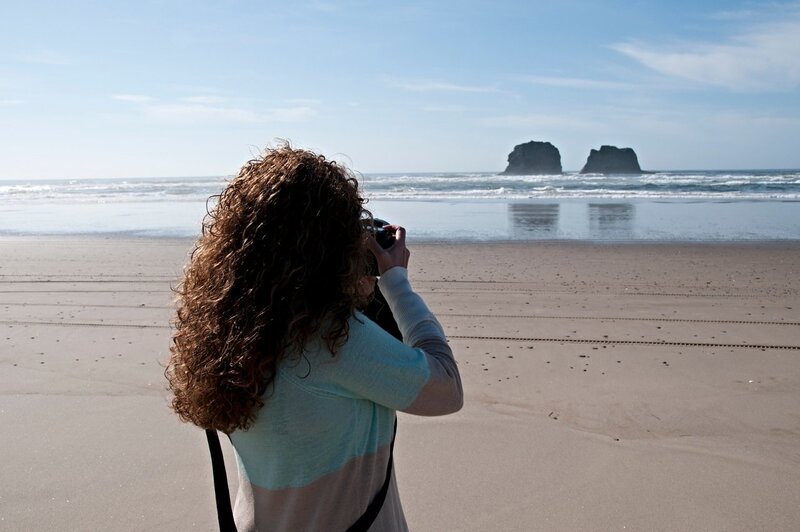 A person with curly hair wearing a light-colored jacket is standing on a sandy beach, facing the ocean and taking a photo. The sky is clear, and two large rock formations are visible in the water in the distance.