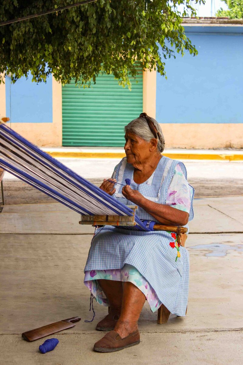 Elderly woman weaving on a traditional backstrap loom in Oaxaca, seated under the shade of a tree with colorful buildings in the background ©Stephanie Dosch | theViatrix Mexico Private Luxury Travel
