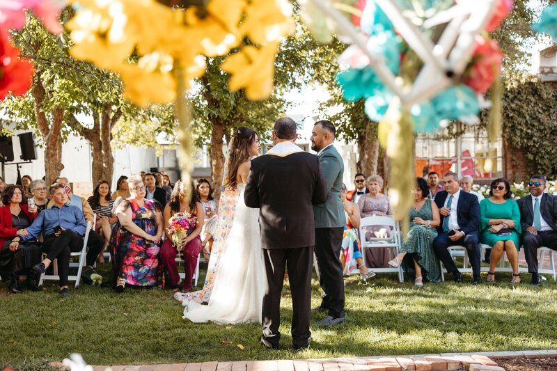 A bride and groom stand with their officiant in front of a colorfully dressed crowd