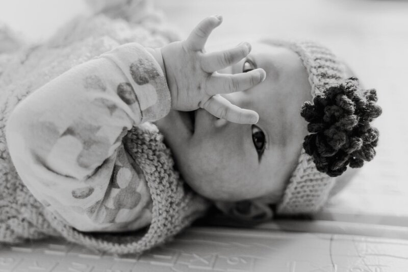 Black-and-white close-up of a baby wearing a knitted hat with a flower detail, sucking on their hand while lying down.