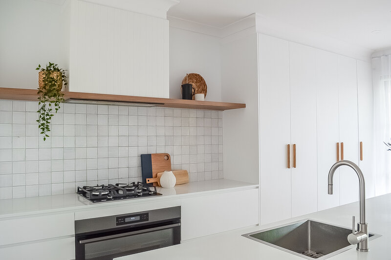 Modern kitchen with white cabinets, gas stove, wooden shelf with decor, and silver faucet on island sink, crafted by a bespoke interior design team in Newcastle NSW.